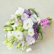 Shaped by experienced hands in Stanmore and set upon a subtly woven beige backdrop, this modern bouquet overflows with tones of creamy white, pastel lavender, and fresh spring green. Ruffled Lisianthus blossoms, plump and luminous, serve as the focal point, offering a soft, inviting texture that echoes the tranquillity of Stanmore's leafy avenues. Around them, unopened buds in shades from lime to ivory hint at new beginnings, joined by clouds of delicate lavender-pink stock flowers that imbue the bouquet with gentle colour and understated fragrance. Spiky globes of deep purple allium create dramatic contrast, their intricate star-shaped petals adding a sculptural element suggestive of artistic displays at local gatherings. Every detail is carefully accented with loops of bright bear grass and feathery lime greens, creating movement and depth, as if tracing the meandering paths of Stanmore's woodlands. The arrangement is finished with a vibrant fuchsia satin ribbon and a crisscross of lustrous white pearls-details that blend tradition and contemporary style, making this bouquet ideal for a wedding, bridal shower, or unique event held in Stanmore's welcoming community. Enveloped in soft light, the palette and textures evoke a spirit of celebration, artistry, and the natural joys cherished in this corner of North West London.
