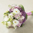 A carefully hand-tied bouquet lies horizontally on a pale beige cloth that resembles smooth burlap or linen, its neutral tone allowing the flowers to glow softly, as though lit by gentle afternoon light coming through a Stanmore bay window. At the heart of the design, large white lisianthus blooms open like silky rosettes, each petal curving inward to reveal a soft yellow-green centre, while half-closed buds add freshness and dimension. Surrounding them, pastel clusters of stock in blush pink and light mauve create a cushion of ruffled texture, their many small blossoms giving the impression of clouds drifting through the arrangement. Piercing this softness are distinctive spikes of purple filler flowers, similar to ornamental allium, with tiny star-shaped petals that form rounded bursts of vibrant lavender-purple, providing structure and a playful, almost firework-like rhythm. Slender green blades of foliage, possibly bear grass, sweep and loop through the bouquet, especially along the right side, tracing elegant curves that make the composition feel fluid and alive, like a walk through Canons Park in late spring. The stems are neatly aligned and wrapped in a vivid fuchsia satin ribbon, tightly wound for comfort in the hand. Over this, a fine strand of luminous white pearls is pinned in place, lending a classic, bridal elegance. The overall mood is romantic yet polished, ideal for a Stanmore bride seeking something timeless but with a touch of colour, or for marking an anniversary dinner at home near The Broadway. It suggests soft fragrance, cool petals, and the reassuring weight of a thoughtfully designed bouquet created with care by an experienced local florist.