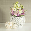 Soft morning light spills across a clean white surface to reveal a lush, rounded floral arrangement in soothing pastels. At the heart of the design, creamy white and pale yellow lisianthus unfurl in generous, velvety rosettes, lifting above clusters of blush-pink stock that add a romantic, fragrant haze. Interspersed among them are small, spiky purple alliums that introduce a playful, architectural note and a delicate contrast of shape. Fresh green foliage fills the negative space, its glossy texture offset by a darker green leaf band wrapped around the bouquet's base, neatly securing the stems and giving the design a finished silhouette. The flowers are housed in a unique spherical vase made of interlaced white branches, a sculptural lattice that feels both airy and crafted, allowing glimpses of stems and water while maintaining a crisp, contemporary profile. Nearby, two open-weave wooden or rattan spheres rest casually to the left, one noticeably larger, repeating the woven motif and adding a touch of natural, tactile charm. The backdrop is a softly textured beige or greige wall-perhaps a grasscloth or linen finish-blurred just enough to let the blooms breathe visually. The overall mood is one of quiet refinement and approachable elegance, a bouquet that would sit comfortably in a Stanmore sitting room or act as a thoughtful gift for a friend returning from a walk through Canons Park. The scent would be light and green, and the balance of softness and structure makes this arrangement equally suited to intimate celebrations or calm, restorative moments.