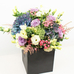 Expertly crafted in Stanmore, this lush floral arrangement showcases an abundance of freshly gathered hydrangeas in deep indigo and periwinkle blue, layered with lavender and blush pink roses that nestle gently together in a contemporary matte black square box. Creamy white and pale yellow lisianthus spill upward, their ruffled trumpets and soft buds suggesting the lightness of spring breezes over Bentley Priory Nature Reserve. Vivid, brainy tufts of lime green filler flowers-likely Green Trick dianthus-peek through, providing playful contrast amidst sprays of delicate, pastel pink astilbe. The design is enriched by an artist's palette of foliage: glossy dark leaves frame lighter, brighter greens, ensuring the composition remains fresh, vibrant, and full from every angle. The design's shape and palette echo the sophisticated, breezy style favoured in Stanmore's homes and businesses, while the sleek matte black container nods to both modernity and timeless luxury. Illuminated against a clean white backdrop, the bouquet positively glows, making it an ideal centerpiece for celebrations, client gifts, or serene sympathy tributes delivered throughout Stanmore. Every bloom whispers a message of thoughtfulness, artistry, and local pride.