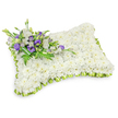 A tender, pillow-shaped sympathy tribute formed from hundreds of small, pristine white chrysanthemum heads, each bloom packed close to create a cloud-like, plush surface that reads as quiet and reverent. The rectangular cushion has gently rounded corners and a slightly raised, puffy profile, the effect of a soft resting pillow rendered entirely in living petals. Around its outer edge a pale lime-green ribbon is pleated into neat overlapping loops, a delicate scalloped frame that lifts the white expanse and gives the composition a fresh, hopeful accent. Diagonally across the upper-left corner rests a composed cluster of accent flowers and foliage: soft white lisianthus with their layered, silky petals sit alongside deep purple lisianthus-some in tight buds-whose richer tones punctuate the otherwise pure, neutral field with dignity and warmth. Feathery fern fronds and broad, dark-green leaves weave through the spray, adding contrast, texture, and the subtle scent of fresh greenery. A broader band of deeper green leaf wraps the base of the small bouquet, neatly binding the stems and lending an understated, crafted finish. The tribute is shown against a clean white background with a faint shadow beneath it, which emphasizes its three-dimensional form and the careful handiwork of a sympathetic arrangement. The overall mood is one of serene comfort and respectful remembrance-ideal for graveside placement or a memorial service-and the sensory impression is of soft petals, gentle green freshness, and a composed stillness meant to console those gathered in quiet memory.
