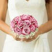 In this image, the story of a Stanmore wedding is delicately unveiled: a bride, serene and jubilant, nestles a bouquet of pink roses against her heart. Each bloom reveals a carefully crafted gradient-soft pastel pink centers expanding outward into vibrant magenta leaves-recalling sunrise hues over Stanmore's rolling hills. Sprigs of baby's breath weave among the roses, casting a gauzy, veil-like softness across the ensemble, while the bride's warm, gently tanned hands hold the bouquet with gentle reverence. Her nails gleam softly, and her sleeveless dress-a vision of embroidered lace and an elegantly gathered sash-completes the portrait. The neutral, blurred background ensures that every nuanced textural detail, from the silken rose petals to the froth of gypsophila, rises to prominence. Intended for the most heartfelt of Stanmore celebrations and prepared with the artistry and warmth local florists are cherished for, this bouquet tells a tale of hope, joy, and everlasting love-where every fibre, petal, and shimmer is an ode to a moment that will never be forgotten.