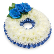 Photographed from a gentle overhead angle against a clean white backdrop, this circular sympathy wreath reads as a study in quiet contrasts and serene textures. The main body is formed from a dense, continuous ring of pristine white chrysanthemums - their feathery, layered petals build a soft, slightly domed surface that appears almost cloud-like and invites a gentle, restorative stillness. Nestled at the upper-left quadrant are three deeply saturated blue roses; their velvety, spiralled petals catch the light with a subtle sheen and stand in solemn counterpoint to the snow-white chrysanthemum field. Rich, dark green foliage peeks out beneath the blue roses, adding natural depth and a tactile fullness that balances the composition. A pleated satin ribbon in matching cobalt traces both the outer and inner rims of the chrysanthemum ring, its glossy folds catching highlights and shadow to frame the design with refined dignity. The overall feel is calming and reverent - a crafted tribute that speaks of remembrance, consolation, and respectful presence. You can almost sense the faint, gentle scent of chrysanthemums mingling with the deeper, subdued perfume of the roses; the soft shadow beneath the wreath gives it weight and presence on the white surface. As a local florist familiar with Stanmore's quiet corners - from Stanmore Broadway to Canons Park - I picture this arrangement placed respectfully at a memorial or chapel, where its restrained palette of white and blue will comfort guests and honour lost connections with grace and tenderness.