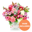 A radiant mixed bouquet of pink blooms sits in a neat square glass vase, photographed against a brilliant white background that showcases every petal and leaf with clarity. At the forefront are large pink roses, some saturated in rich fuchsia and magenta tones and others washed in soft blush and pastel hues, creating a romantic gradient of colour. Interwoven with them are plush carnations in coral and peach-pink, their deeply ruffled petals adding volume and a tactile softness that suggests gentle movement. Sprinkled throughout the bouquet are small daisy-like chrysanthemums in lavender and muted purple shades, each with a striking lime-green centre that pops against the surrounding pinks. Creamy white flowers peek out between these stronger tones, providing a sense of lightness and balance. Embracing the blooms is a mix of greenery: rounded eucalyptus leaves in silvery blue-green, darker green foliage providing structure, and a few discreet reddish-brown leaves lending a subtle, cosy warmth. The stems are gathered into a clear square glass vase whose crisp lines give the arrangement a contemporary edge. Around the base of the vase, a wide white band is wrapped securely, decorated with repeating small red hearts and the message all my love written in flowing script, turning the container into part of the sentiment. In the lower right-hand corner of the photo, an orange circular icon with bold white capital letters announces VASE INCLUDED, highlighting that it arrives ready to display. The bouquet evokes the feeling of a lovingly prepared gift from a Stanmore florist, perfect for brightening a home near Stanmore station, along Marsh Lane, or by the leafy surroundings of Canons Park with a burst of colour and affection.